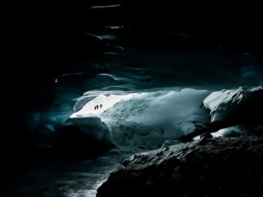 Intérieur d'une grotte de glace contrastant avec la lumière du jour et la présence de trois silhouettes humaines.