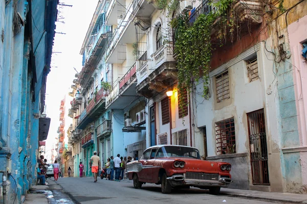 Scène de rue dans un quartier historique avec une voiture ancienne rouge et des passants.