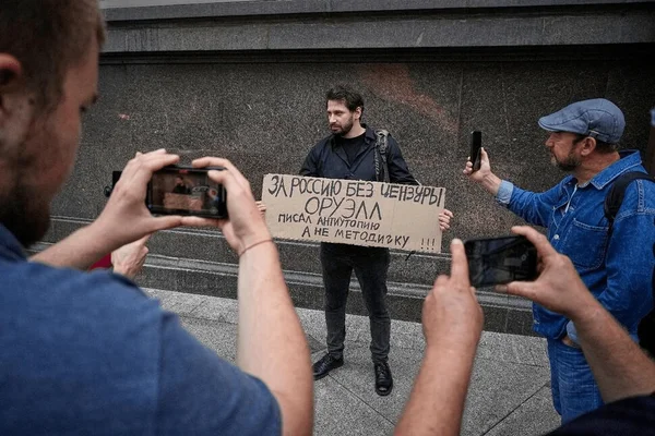 Un homme brandissant un panneau en carton avec du texte russe, encadré par des personnes prenant des photos avec leurs smartphones.