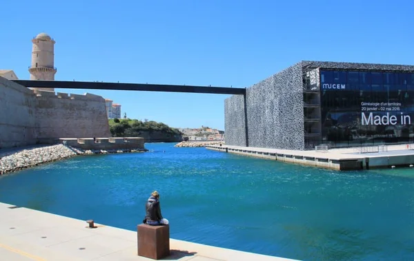Le fort Saint-Jean et le Mucem vus du bord de l'eau, avec un passant assis sur un piédestal.