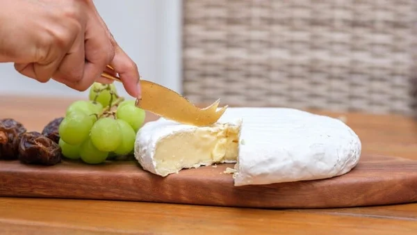 Le geste de couper un camembert avec un couteau doré, accompagné de fruits secs.