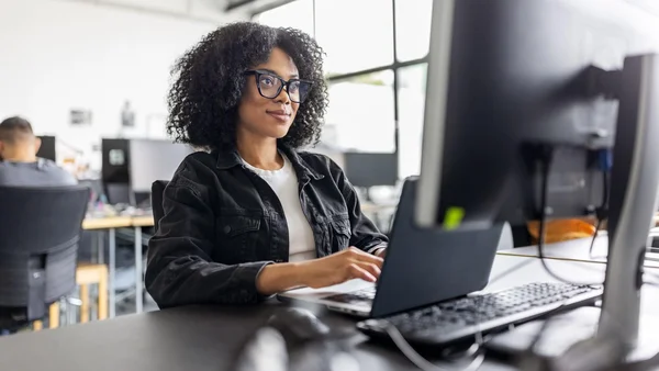 Une femme aux cheveux bouclés et portant des lunettes travaille sur un ordinateur portable dans un environnement de bureaux.