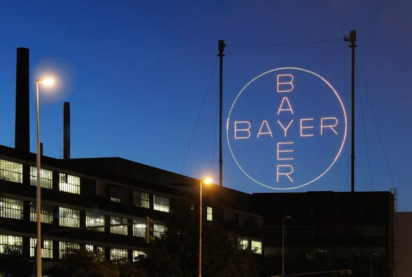 Le logo circulaire Bayer illuminé la nuit devant un bâtiment aux fenêtres allumées.