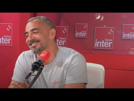 Julien Mairesse assis en studio à France Inter, tenant le micro rouge de la station sur le fond rouge emblématique.