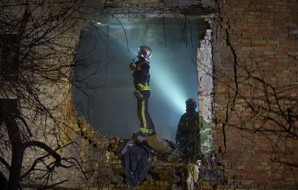 Des pompiers sur les ruines d'un bâtiment en briques éclairé par des projecteurs la nuit.