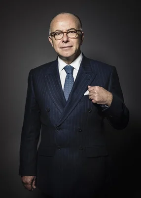 Bernard Cazeneuve pose en costume avec une cravate bleue sur fond sombre.