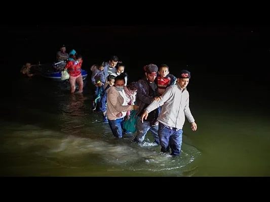 Des migrants, adultes et enfants, progressant dans l'eau de nuit vers la rive, certains étant aidés, un bateau en arrière-plan.