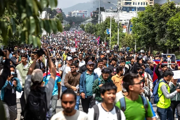 Un homme en gilet orange et topi traditionnel, les mains jointes, entouré d'autres personnes lors d'un rassemblement extérieur.