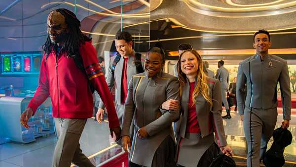 Cinq acteurs en uniformes gris et rouges se déplaçant dans un décor aux courbes et lumières colorées.