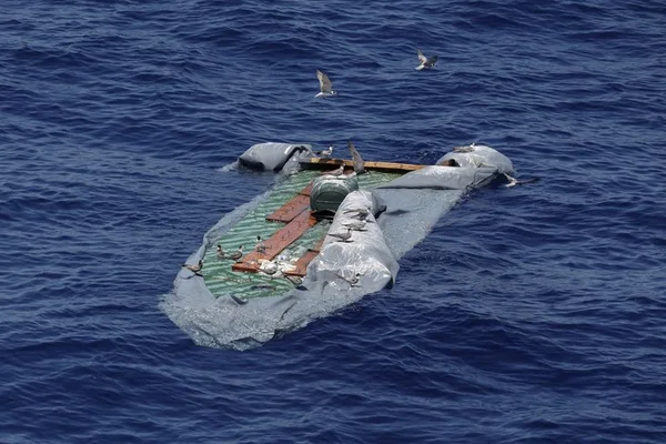 Les débris d'une embarcation migrant partiellement submergée flottant en mer, avec des oiseaux perchés sur la structure.