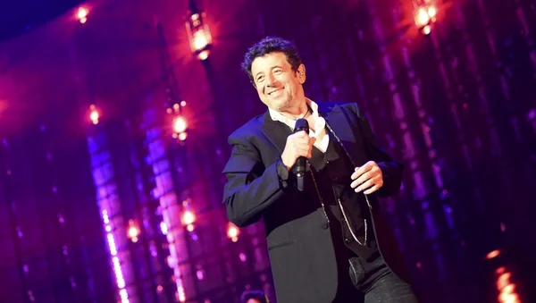 Patrick Bruel chante sur scène avec un micro, sous un éclairage violet et rouge.