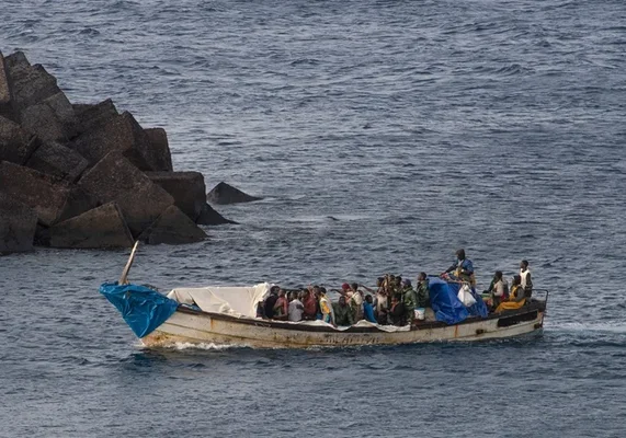 Un bateau surchargé de migrants naviguant près d'une digue rocheuse, avec une bâche bleue recouvrant partiellement le navire.