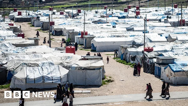Quatre femmes en vêtements longs et sombres debout près de tentes bleues et d'une étendage.