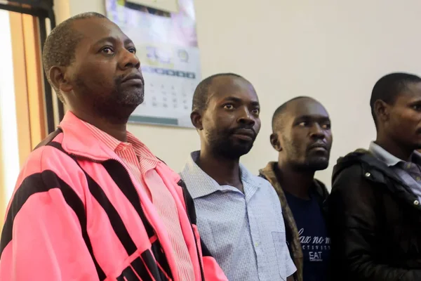 Quatre hommes, dont le pasteur Paul Mackenzie en veste rose, posent devant un mur intérieur avec un calendrier.