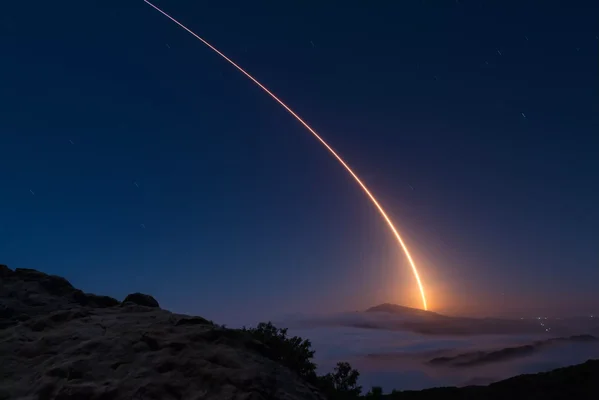 Une traînée lumineuse courbe s'élève dans le ciel étoilé au-dessus d'un terrain rocheux et brumeux.