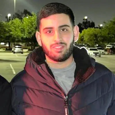 Un homme portant une veste de couleur sombre et une barbe se tient dans un parking la nuit