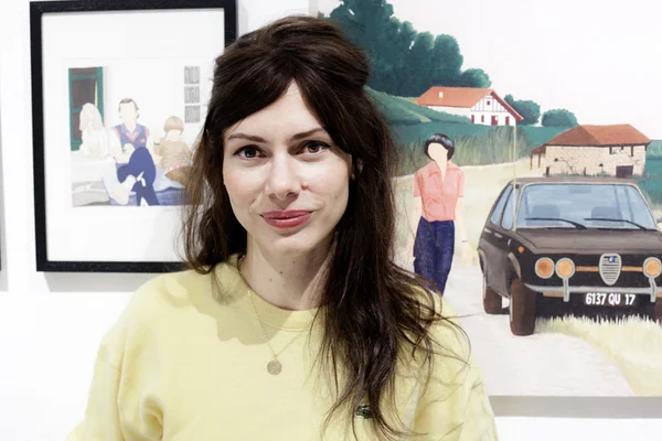 Femme en sweatshirt jaune se tenant devant des peintures, dont une scène rurale figurant une voiture.