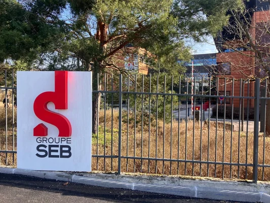 Panneau avec le logo et l'inscription 'GROUPE SEB' devant une clôture métallique et un bâtiment.