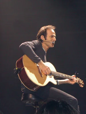 Jean-Jacques Goldman assis jouant de la guitare acoustique sur une scène sombre.