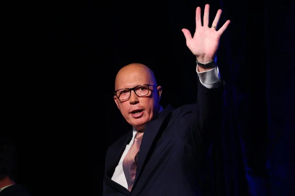 Un homme chauve en costume sombre, portant des lunettes et une cravate à motifs, lève la main en signe d'adieu sur fond sombre.