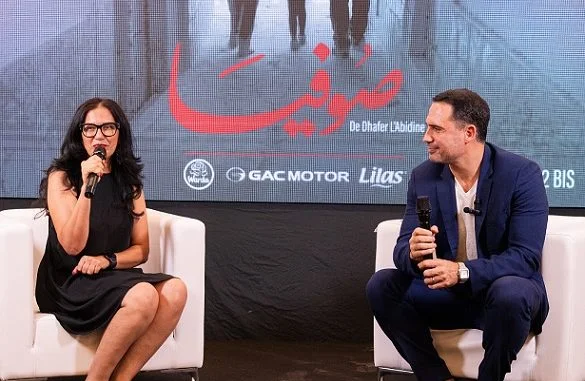 Deux intervenants lors d'un événement de présentation du film Sofia avec des sponsors visibles.