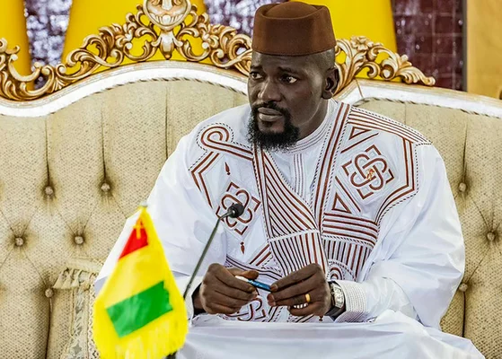 Une figure politique en tenue blanche fait un signe à la foule depuis un véhicule.