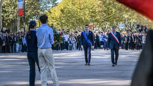 Deux élus en écharpes tricolores défilant avec des fleurs lors d'une cérémonie municipale en extérieur.