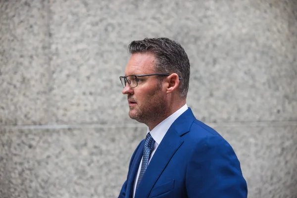 Trevor Milton en costume bleu de profil devant un mur en pierre.