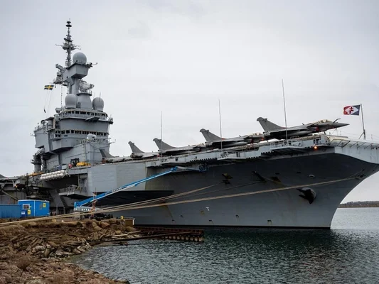 Le porte-avions à quai avec des avions de chasse sur son pont, vu sous un ciel couvert.