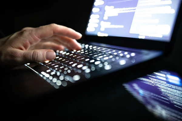 Une main tapant sur un clavier lumineux avec du code affiché sur l'écran d'un ordinateur portable