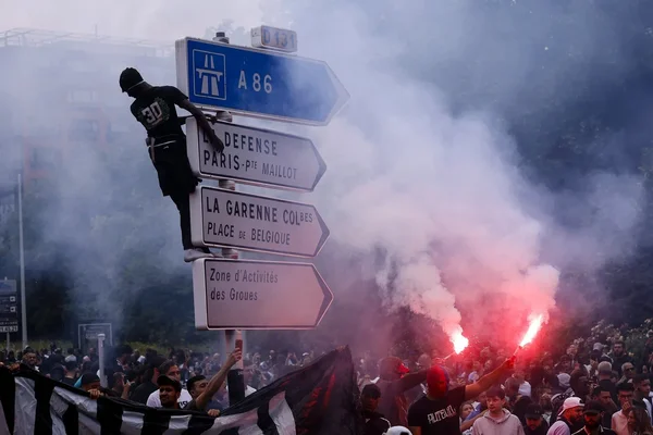 Un groupe au panneau La Défense, dont un individu qui y grimpe, entouré de fumigènes rouges.