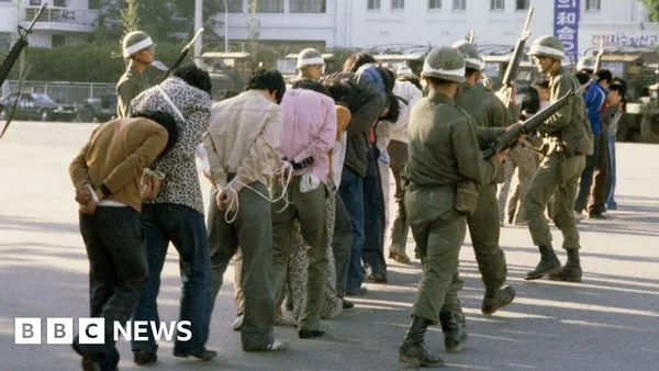 Groupe de civils aux mains liés escortés par des soldats en uniforme militaire.