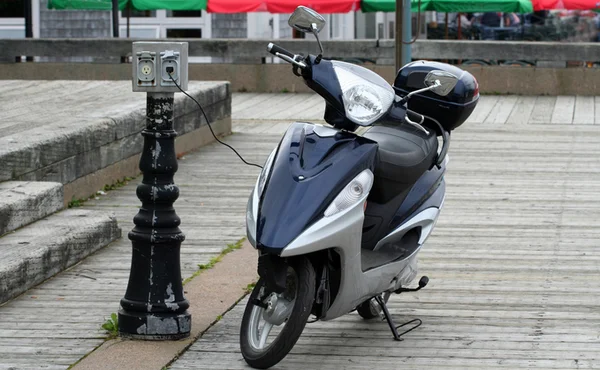 Un scooter électrique bleu et argent garé sur une promenade en bois, branché à une prise électrique.