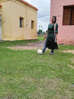 Une femme en tenue décontractée pose avec un ballon de football entre deux bâtiments colorés.