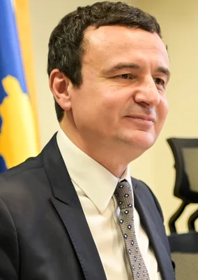 Portrait d'Albin Kurti en costume sombre et cravate à motifs, pris en intérieur avec un drapeau en arrière-plan en 2020.