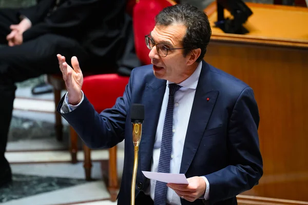 Homme en costume bleu parlant au micro devant un bureau, tenant un document à la main, lors d'une déclaration politique.