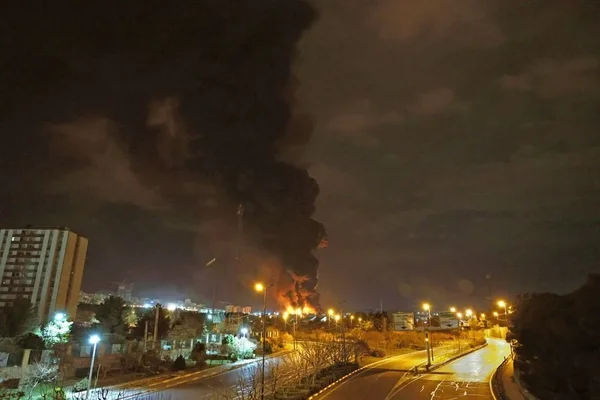 Vue nocturne d'une rue de Téhéran avec un immense panache de fumée noire et des flammes éclairant les bâtiments.