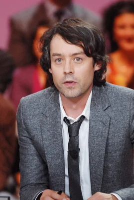 Yann Barthès en costume gris et cravate noire, photographié devant un fond flouté.