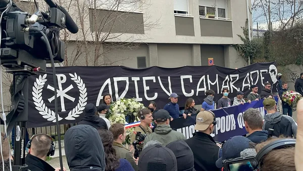 Une bannière 'ADIEU CAMARADE' et des fleurs lors d'un rassemblement.