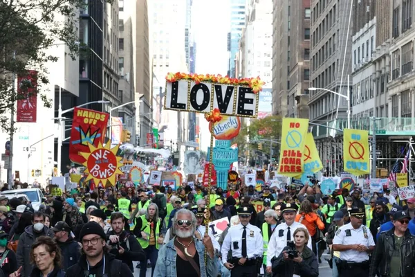 Cortège de manifestants dans une rue avec une pancarte « LOVE» fleurie, encadré par des policiers et photographes.
