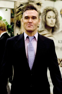 Morrissey photographed in a dark suit, light pink shirt, and light purple tie.