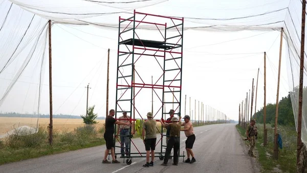 Un groupe déplace un échafaudage métallique sur une route bordée de champs et de filets.