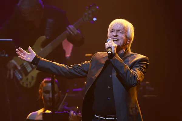 Homme d'âge mûr aux cheveux gris coiffés en arrière, assis derrière un grand piano noir dans un studio d'enregistrement moderne, concentré sur une partition posée sur le pupitre, éclairé par une lumière tamisée bleutée