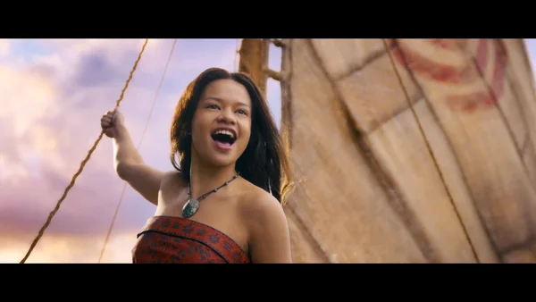 Une actrice en costume de Vaiana sur un voilier, tenant une corde et souriant devant un ciel coloré.