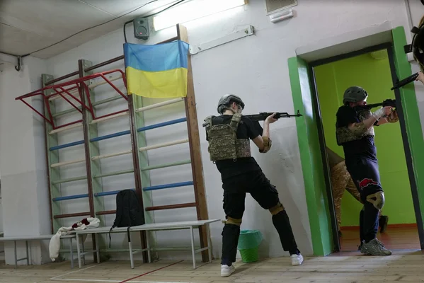 Deux personnes en tenue tactique s'entraînant au maniement des armes dans une salle ornée d'un drapeau ukrainien.