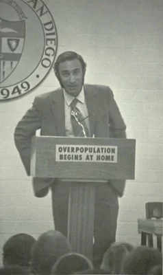 Le biologiste Paul Ehrlich s'exprimant lors d'une conférence à l'Université de San Diego, devant un podium indiquant 'OVERPOPULATION BEGINS AT HOME'.