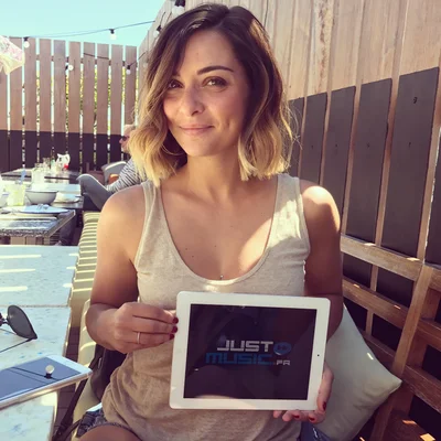 Priscilla Betti assise à une table en extérieur pour une interview, tenant une tablette.