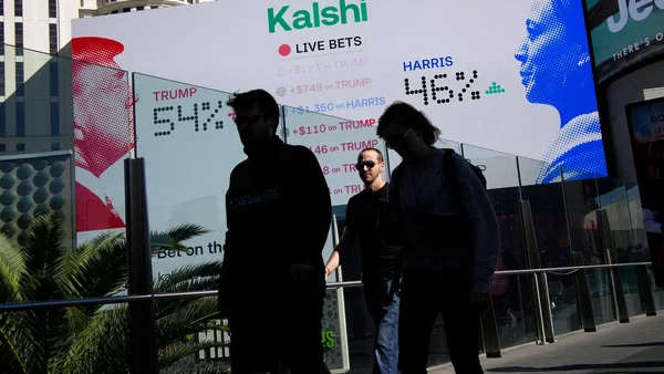 L'image montre une scène de rue avec trois personnes passant devant un grand panneau numérique affichant les cotes de paris en direct pour Trump et Harris, avec Harris à 46 % et Trump à 54 %. Le panneau porte la marque « Kalshi » et inclut des détails de paris comme +749 $ sur Trump et +1 350 $ sur Harris.