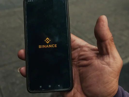 Une main tenant un smartphone affichant le logo Binance, avec un trottoir flou en arrière-plan