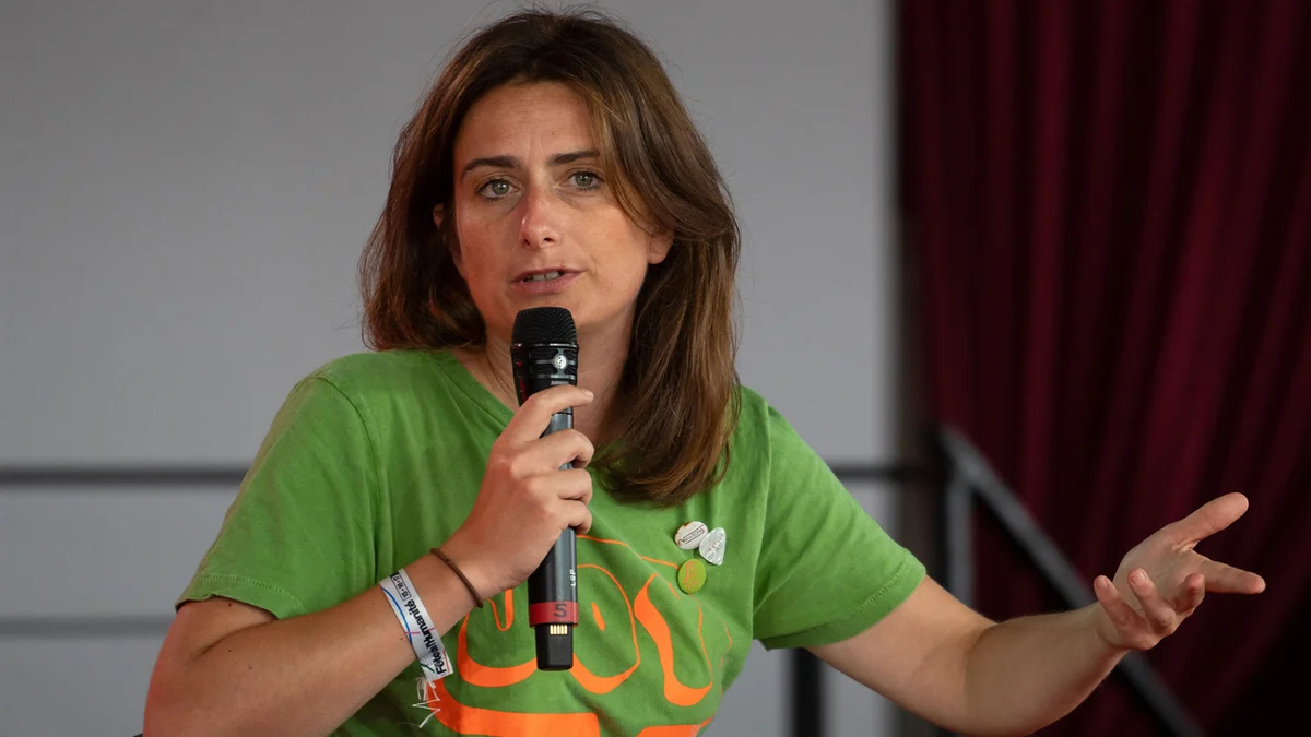 Marine Tondelier en t-shirt vert tenant un micro, devant un décor de rideau rouge.